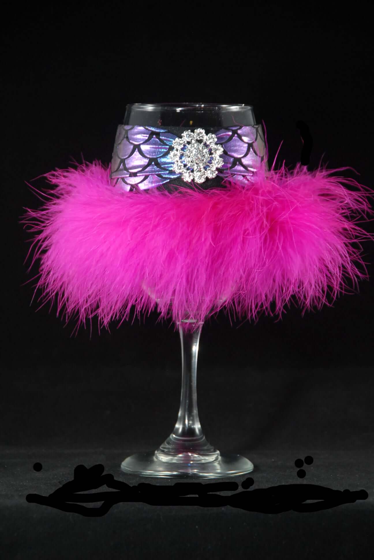 A pink feathered floozie coozie with a circle jewel on a glass, designed to be removable and worn on a wine glass.